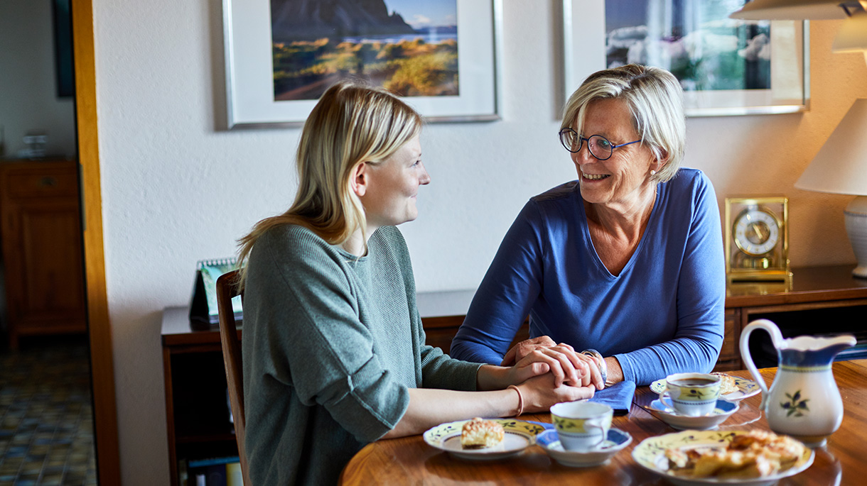 mother and adult daughter sitting at dining table each pastries and drinking tea while holding hands